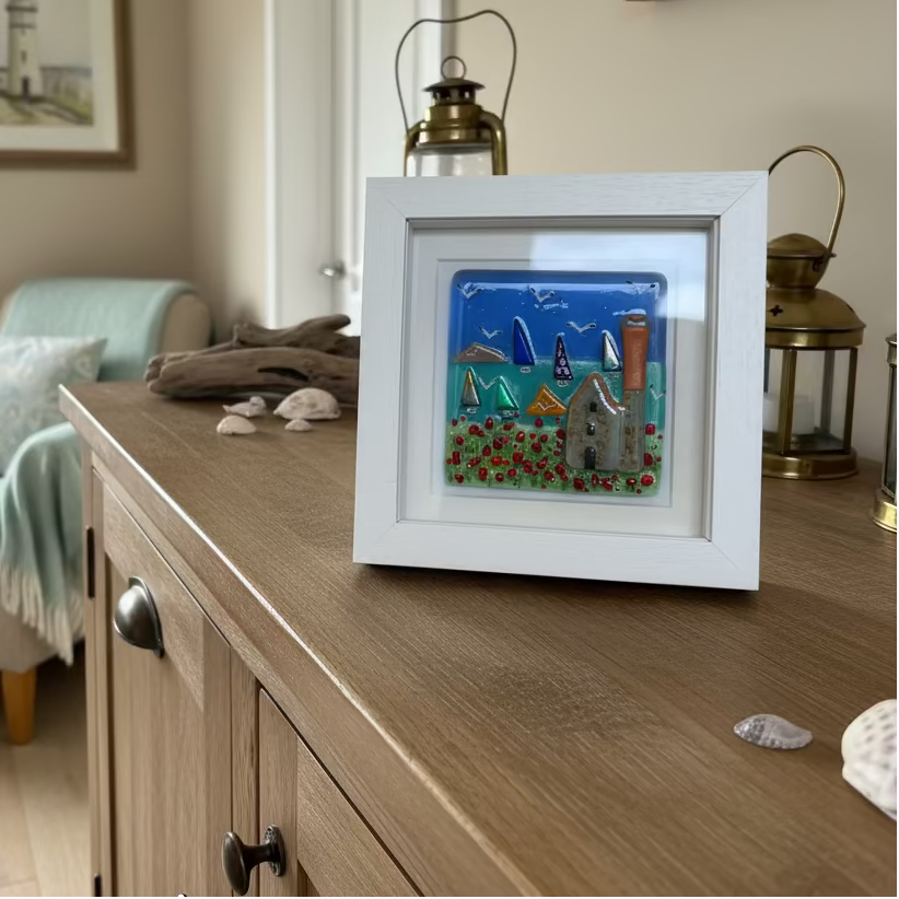 A square white framed glass artwork featuring a colorful coastal scene with sailboats, seagulls, flowers, and a Cornish tin mine, displayed on a wooden tabletop.