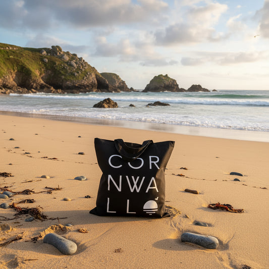 Black Cornwall themed tote bag with white writing sitting on a sandy beach with rocks and sea behind