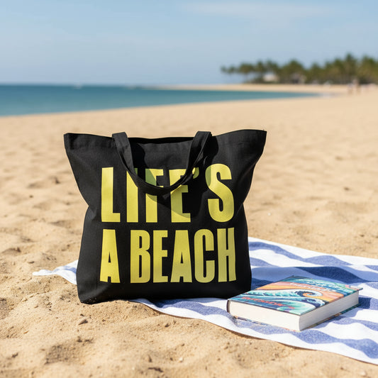 A black tote bag with bright yellow 'LIFE'S A BEACH' text on a sandy beach with a book and towel nearby