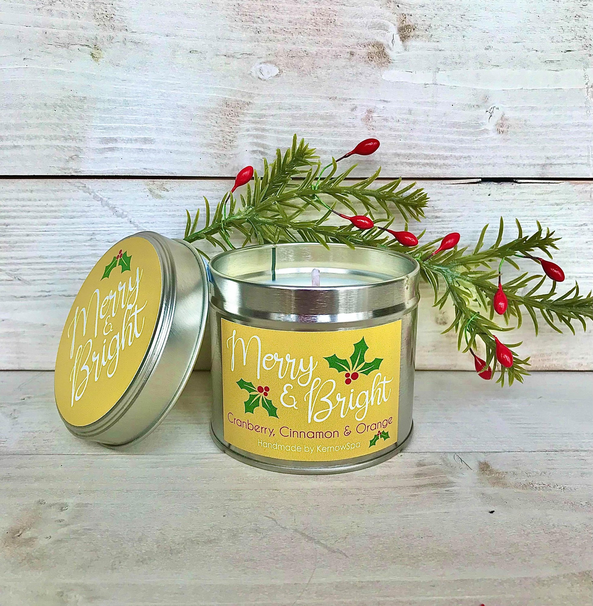 Candle in a metal tin with 'Merry & Bright' label on a wooden surface with holly leaves and berries.