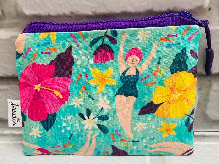 The Wild Swimmer Letterbox Gift Set by Sandy Shores Gift and Home is a vibrant fabric zip pouch showcasing a distinctive design of women in swimsuits amidst flowers and fish on a turquoise background. Ideal as a letterbox gift, it features a purple zipper and the "Jewells" label, set against an elegant brick wall backdrop.