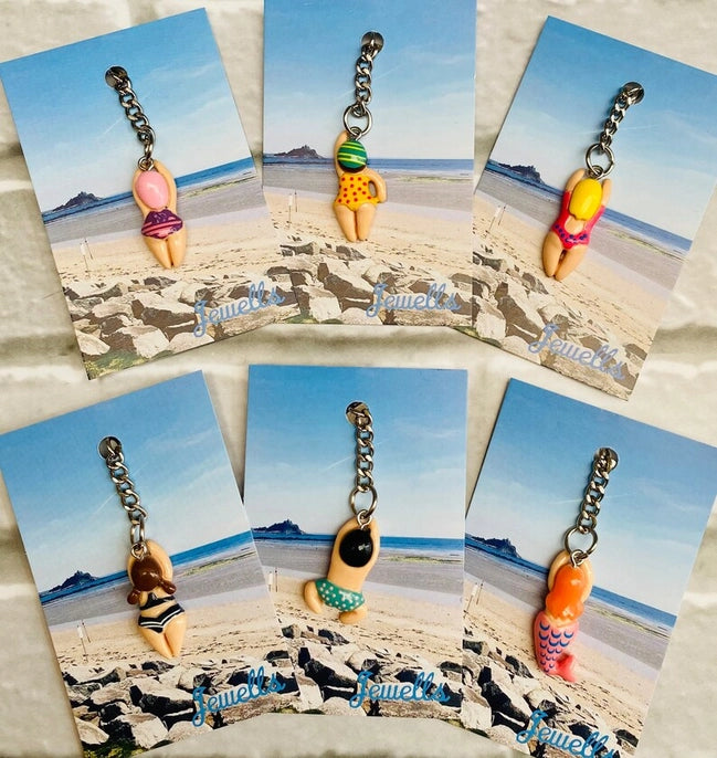 Six vibrant bikini-shaped keychains are displayed on cards featuring a beach and ocean backdrop, each with its own unique design, capturing the essence of a wild swimmer's style. This quirky gift set from Sandy Shores Gift and Home is perfect for sea lovers.