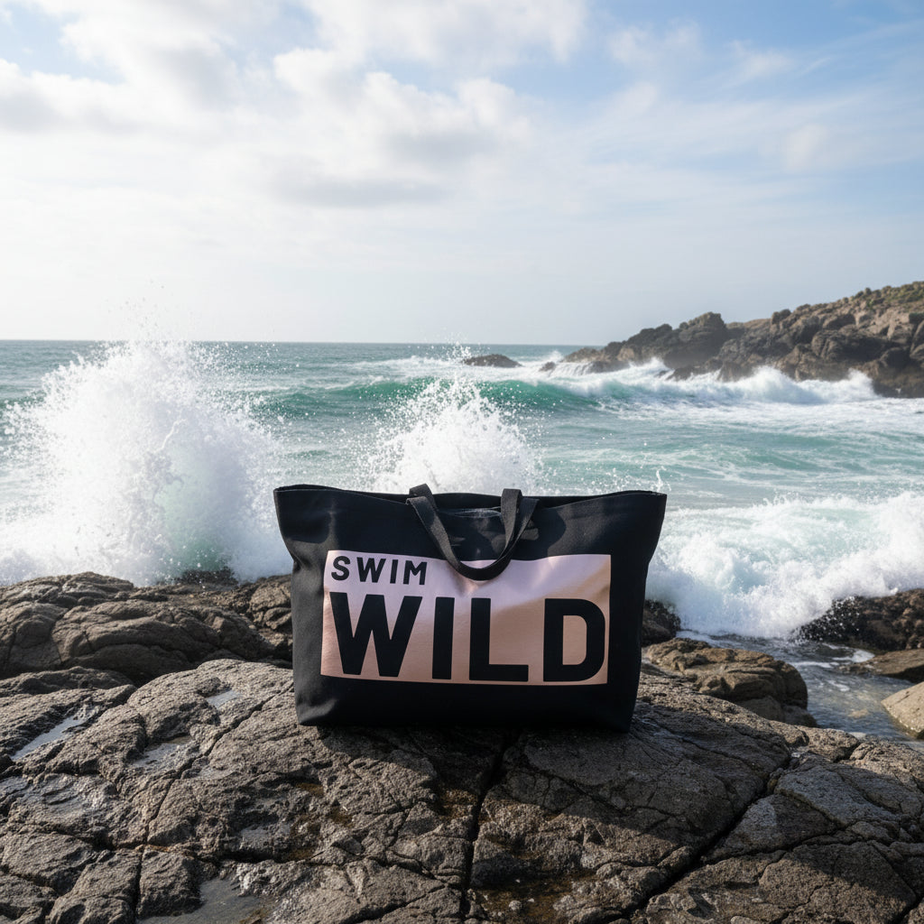 A large black tote bag with a metallic rose gold graphic that reads 'SWIM WILD' in bold black letters on a Cornish beach