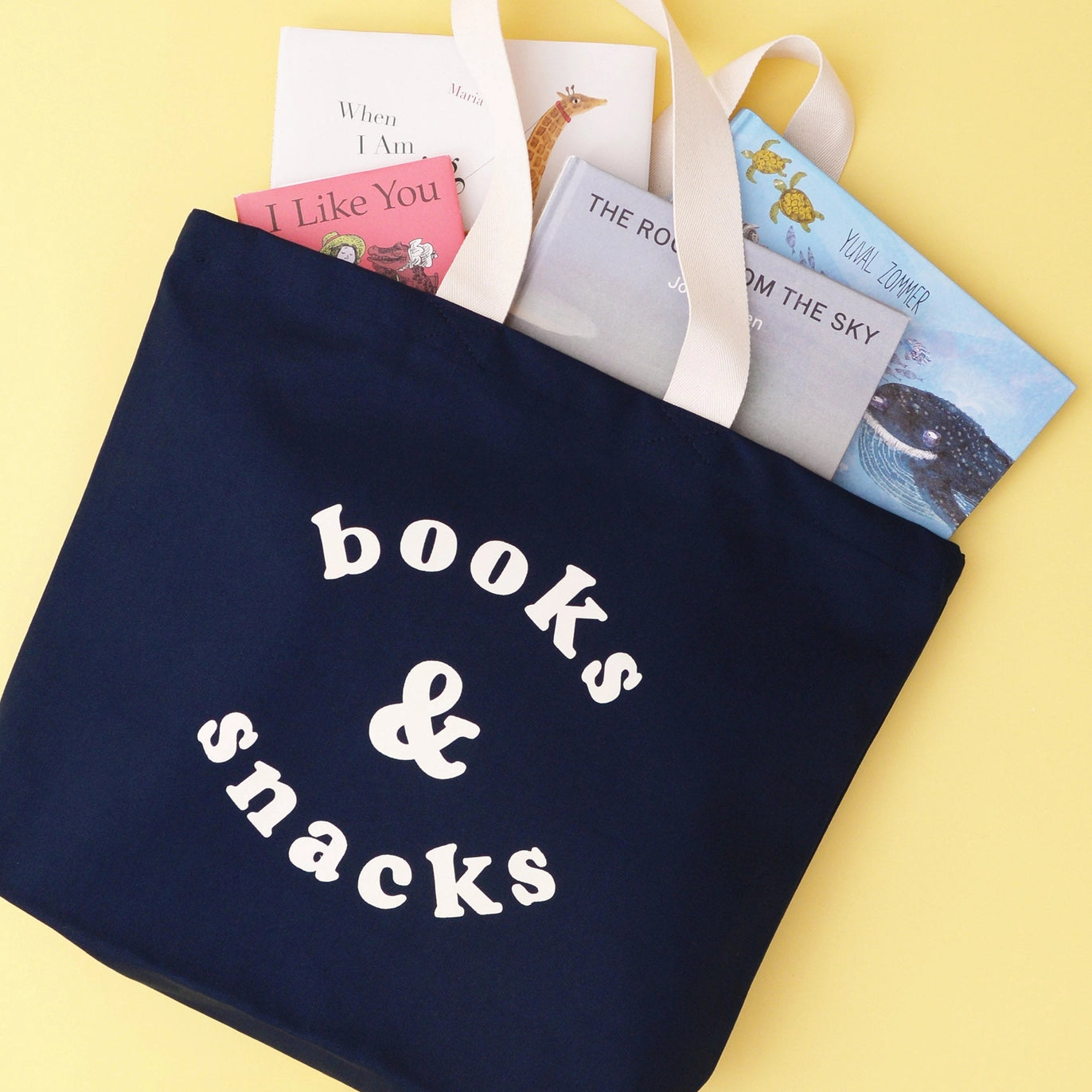 Navy tote bag with 'books & snacks' text, filled with children's books on a yellow background