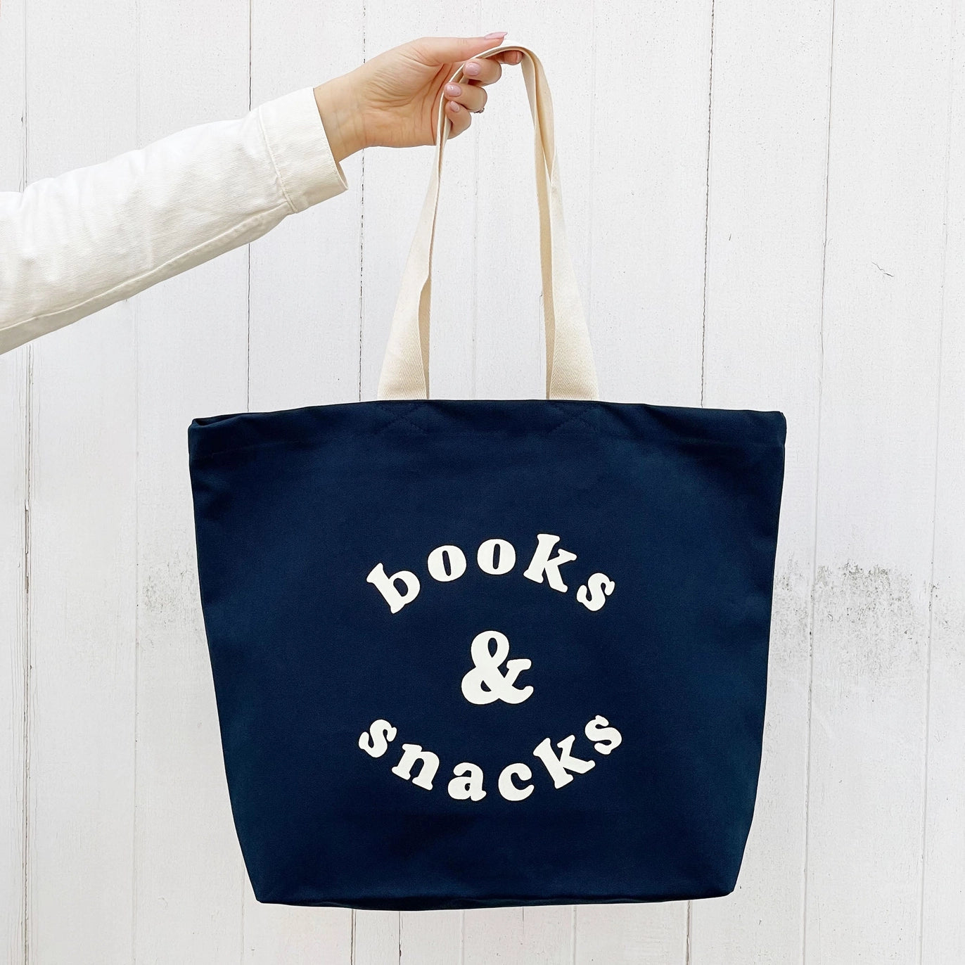 Navy tote bag with 'books & snacks' text held by a person against a white background