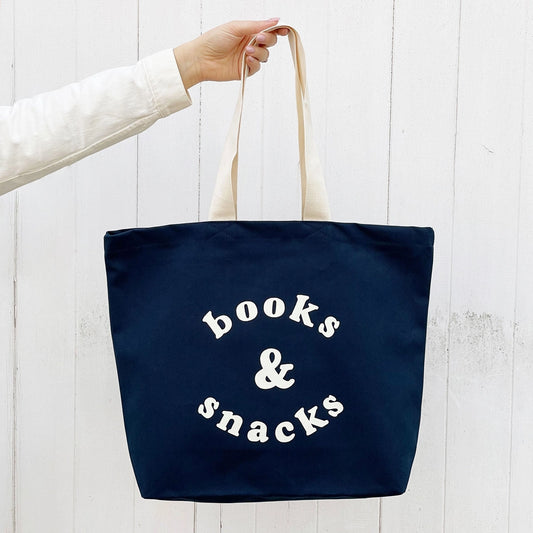 Navy tote bag with 'books & snacks' text held by a person against a white background