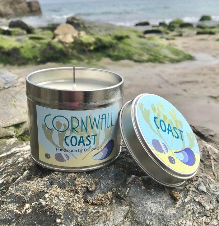 A tin candle labeled "Cornwall Coast" with the lid off, placed on a sandy rock. The background features a beach with rocks and the ocean in sight.