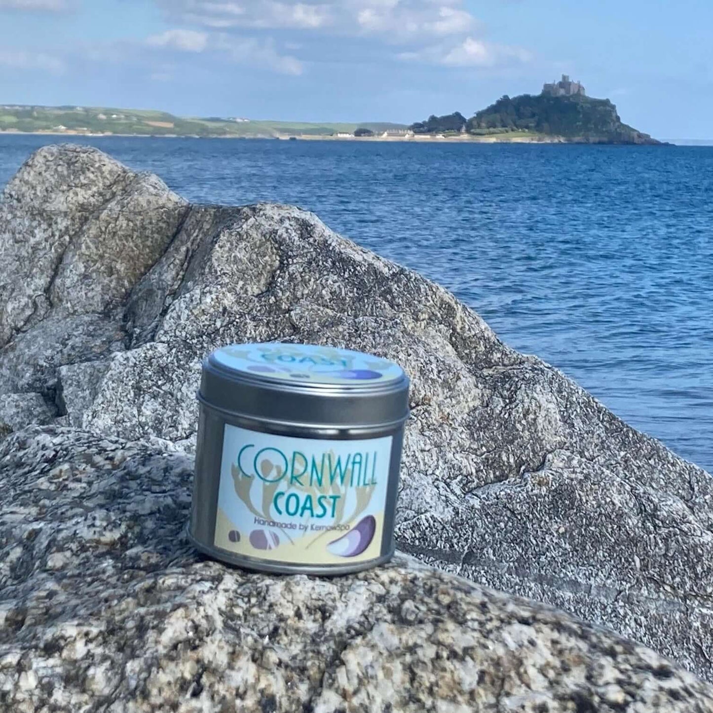 A tin labeled "Cornwall Coast" sits on a rocky sea shore. In the background, there's a large body of water with a distant island. The skies are partly cloudy, and the scene conveys a calm coastal atmosphere.