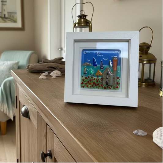 A square white framed glass artwork featuring a colorful coastal scene with sailboats, seagulls, flowers, and a Cornish tin mine, displayed on a wooden tabletop.