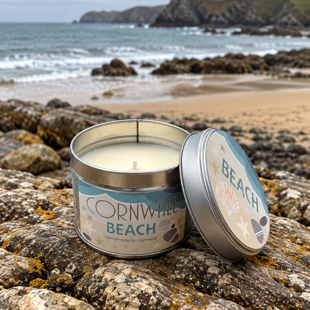 A silver tin candle labeled 'Cornwall Beach' sits on a rocky beach with the ocean and cliffs in the background. The lid rests beside the open tin, and the scene is styled outdoors on natural stone.