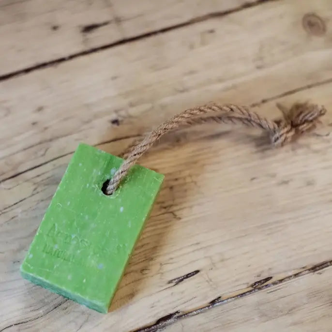 A rectangular green soap with a rustic surface is threaded with a natural brown rope. It is placed on a wooden surface with visible grain patterns.