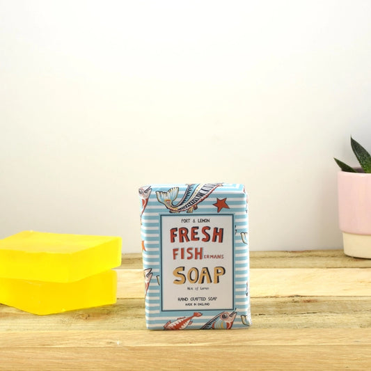 A bar of "Fresh Fishermans Citrus Soap" from Sandy Shores Gift and Home, featuring colorful illustrations, is displayed on a wooden surface. Yellow citrus soap bars are positioned to the left, while a small pink pot with a plant decorates the right side, all set against a plain background that emphasizes its all-natural ingredients.