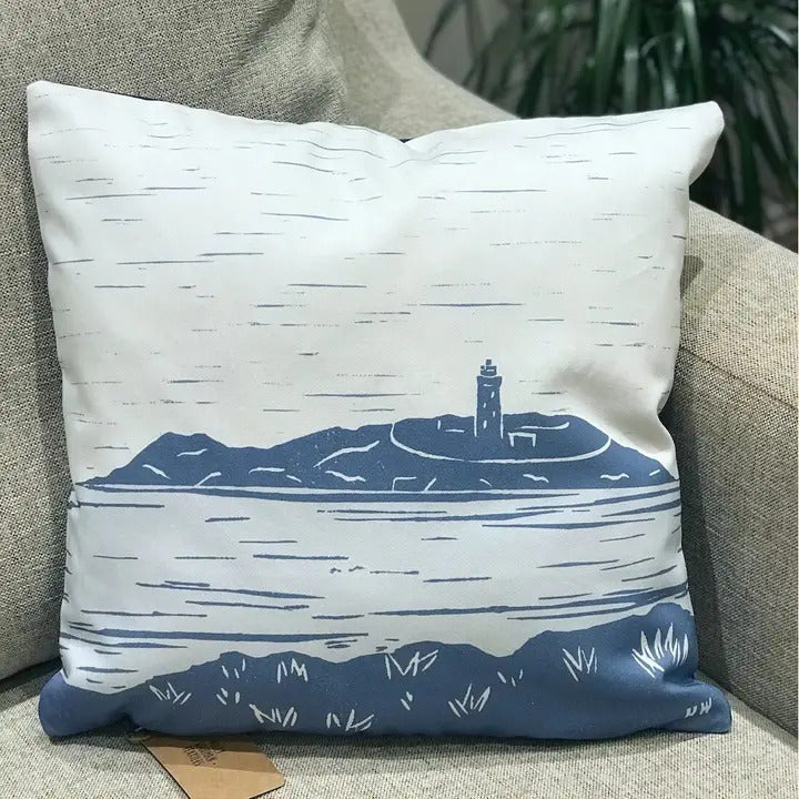 The Godrevy Lighthouse Handmade Luxury Cushion by Cornish Gifts from Sandy Shores showcases a blue and white illustration reminiscent of the lighthouse at St. Michael's Mount in Cornwall. Adorned with a shoreline and ocean waves, this cushion complements any decor, elegantly resting on a beige sofa amidst vibrant green plants.