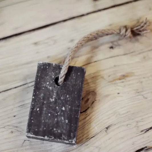 A rectangular bar of soap with a coarse texture is attached to a rustic rope handle. It is placed on a wooden surface, adding a natural and organic aesthetic.