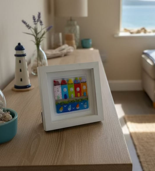 A square white shadow box frame containing colorful, 3D house-shaped glass, with sailing boats in the foreground on a blue background, displayed on a wooden sideboard