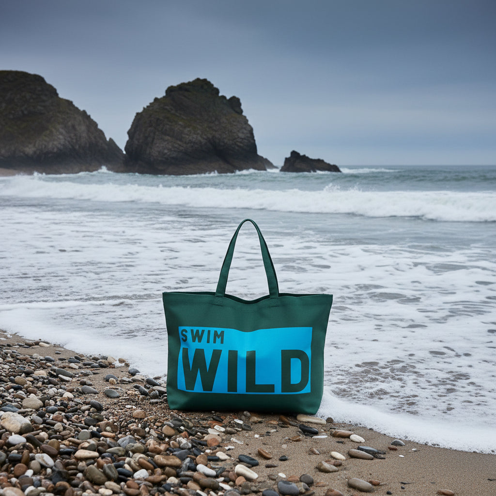 Green tote bag with 'Swim Wild' text on a Cornish beach with the sea behind