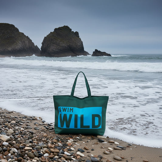 Green tote bag with 'Swim Wild' text on a Cornish beach with the sea behind