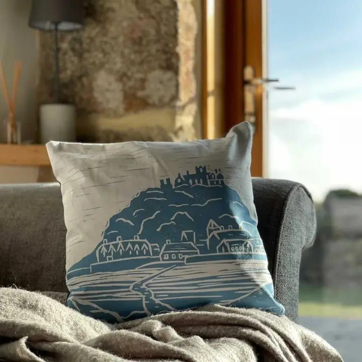 A St Michaels Mount Handmade Luxury Cushion by Cornish Gifts is elegantly displayed on a gray sofa, its blue illustration beautifully offset by the matching tones. A beige blanket adds a touch of warmth to the scene, while sunlight streams through the nearby window, bathing everything in a serene glow.