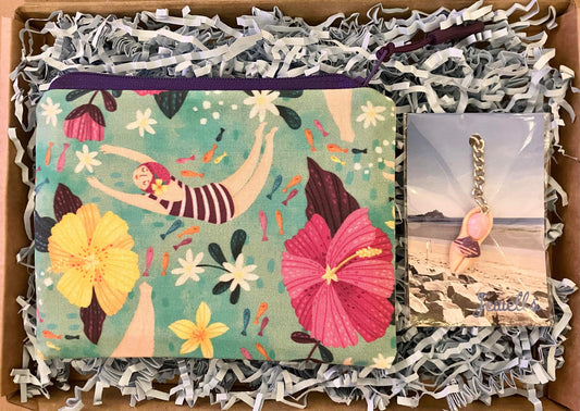 The Wild Swimmer Letterbox Gift Set from Sandy Shores Gift and Home includes a vibrant pouch adorned with a distinctive floral and swimming motif, elegantly presented in a gift box filled with shredded paper. It’s an ideal letterbox gift, complemented by a keychain showcasing a pink seashell set against a beach backdrop.