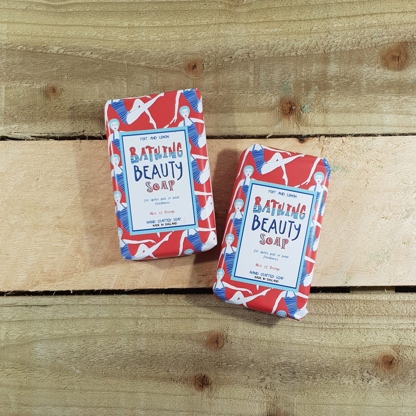 On a wooden surface, there are two bars of Sandy Shores Gift and Home's "Bathing Beauty Citrusy Soap," exuding an invigorating scent. They are wrapped in red and white illustrated paper. This soap is crafted with all-natural ingredients and is labeled as vegan and cruelty-free.