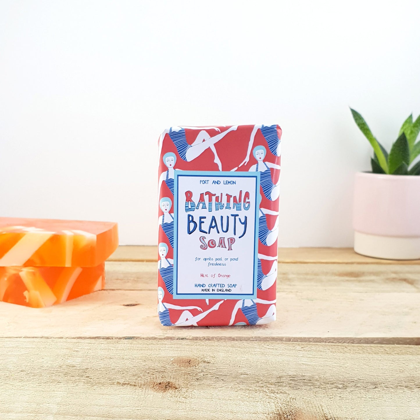 A bar of Sandy Shores Gift and Home's Bathing Beauty Citrusy Soap, adorned with colorful packaging that showcases abstract patterns and figures, sits gracefully on a wooden surface. Exuding an invigorating aroma, it is accompanied by two soap bars sporting orange and white stripes. In the background, a small potted plant enhances the charm of this delightful scene.