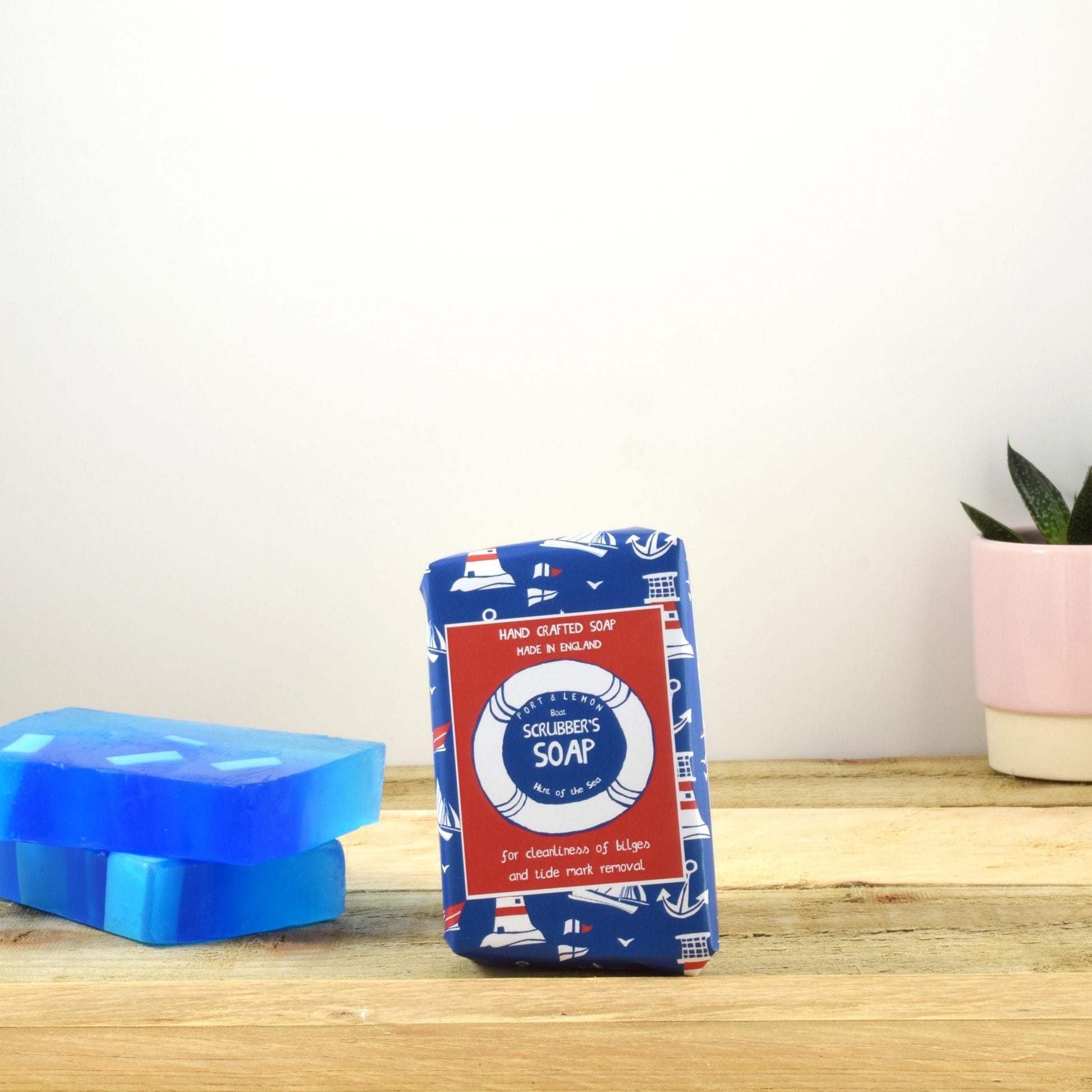 A bar of "Boat Scrubbers Soap Fresh Soap" from Sandy Shores Gift and Home, wrapped in blue nautical-themed paper, rests on a wooden surface. Nearby are two pieces of translucent blue soap, ideal for achieving a sparkling clean. Handcrafted in the UK, a small potted succulent adorns the background on a light wood surface.