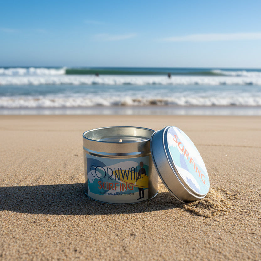 Candle in a metal tin with Cornwall Surfing branding. The candle is on a sandy beach with waves behind