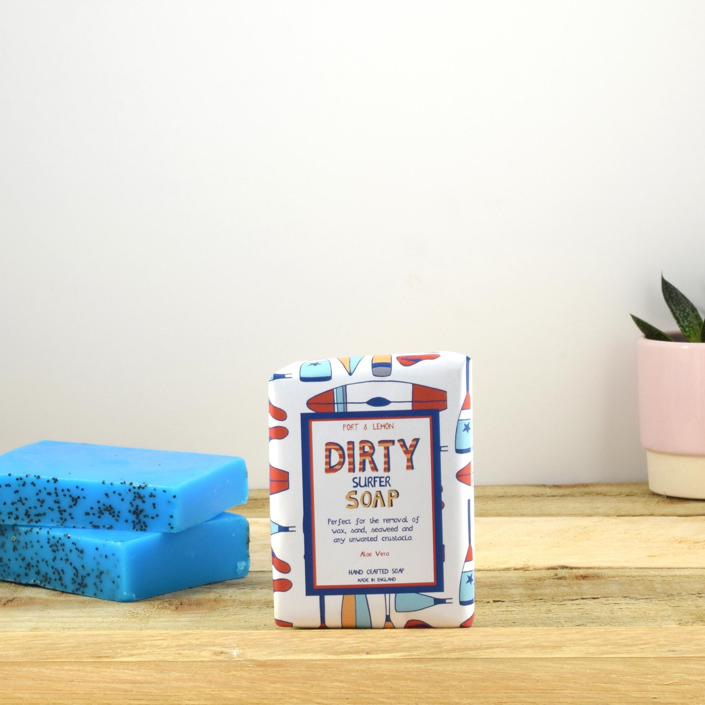 A "Dirty Surfer Soap Bar" by Sandy Shores Gift and Home, meticulously crafted in the UK, is displayed on a wooden surface with eye-catching packaging. Next to it are two blue bars sprinkled with black specks, alongside a small succulent in a pink pot. A light-colored wall accentuates the clean, minimalistic ambiance.