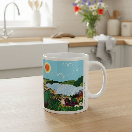 A white ceramic mug featuring a colourful illustration of the eden project under a blue sky, set against a kitchen background.