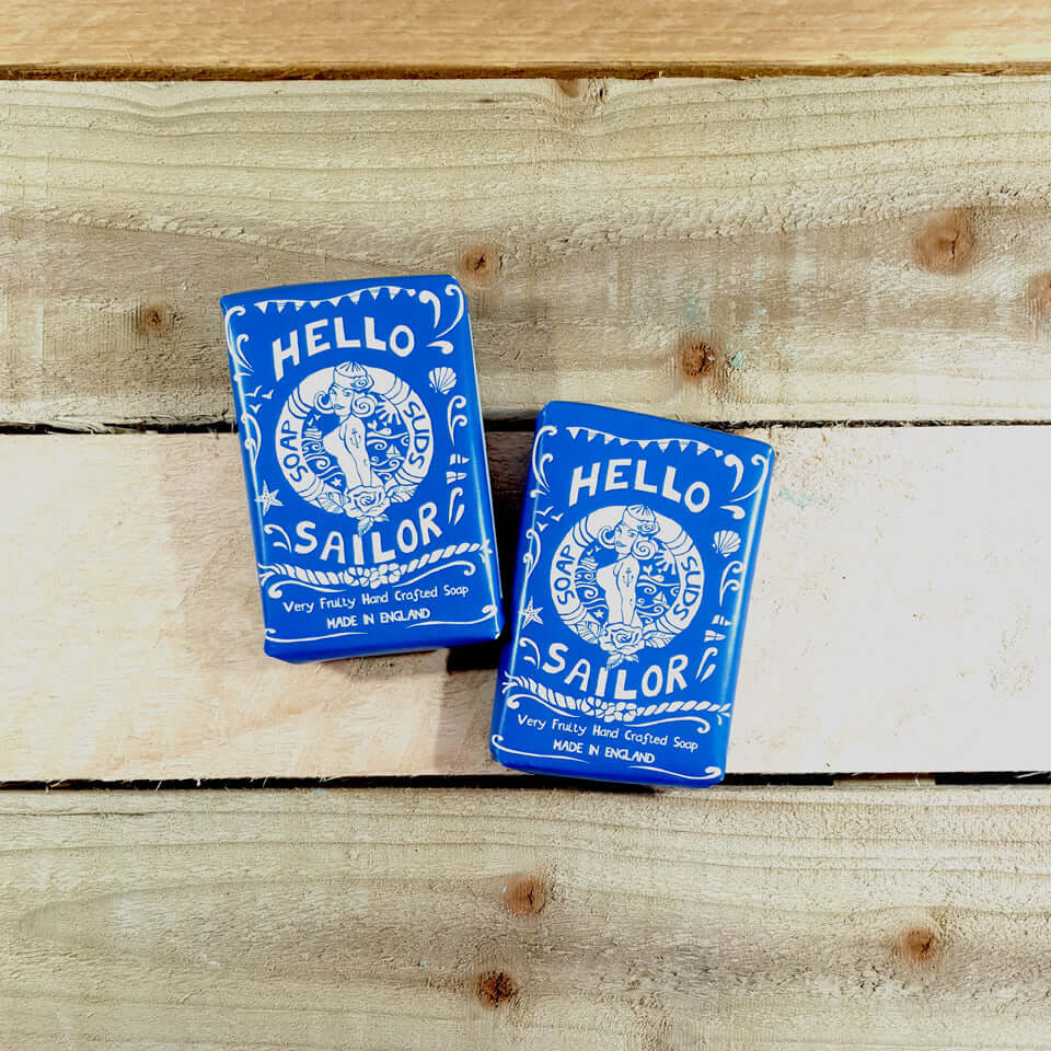 Two blue bars of nautical-themed soap from Sandy Shores Gift and Home feature charming illustrated sailor designs and are labeled "Hello Sailor Citrus Soap" and "Very Fruity Red Clay Soap." Crafted by Bathing Beauty Soap in the UK, these bars rest elegantly on a wooden surface.