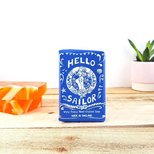 A blue soap box labeled "Hello Sailor Citrus" with decorative white graphics stands on a wooden surface. Two orange and white swirl soaps, bursting with citrus allure, and a small potted plant add charm to the scene in the background.