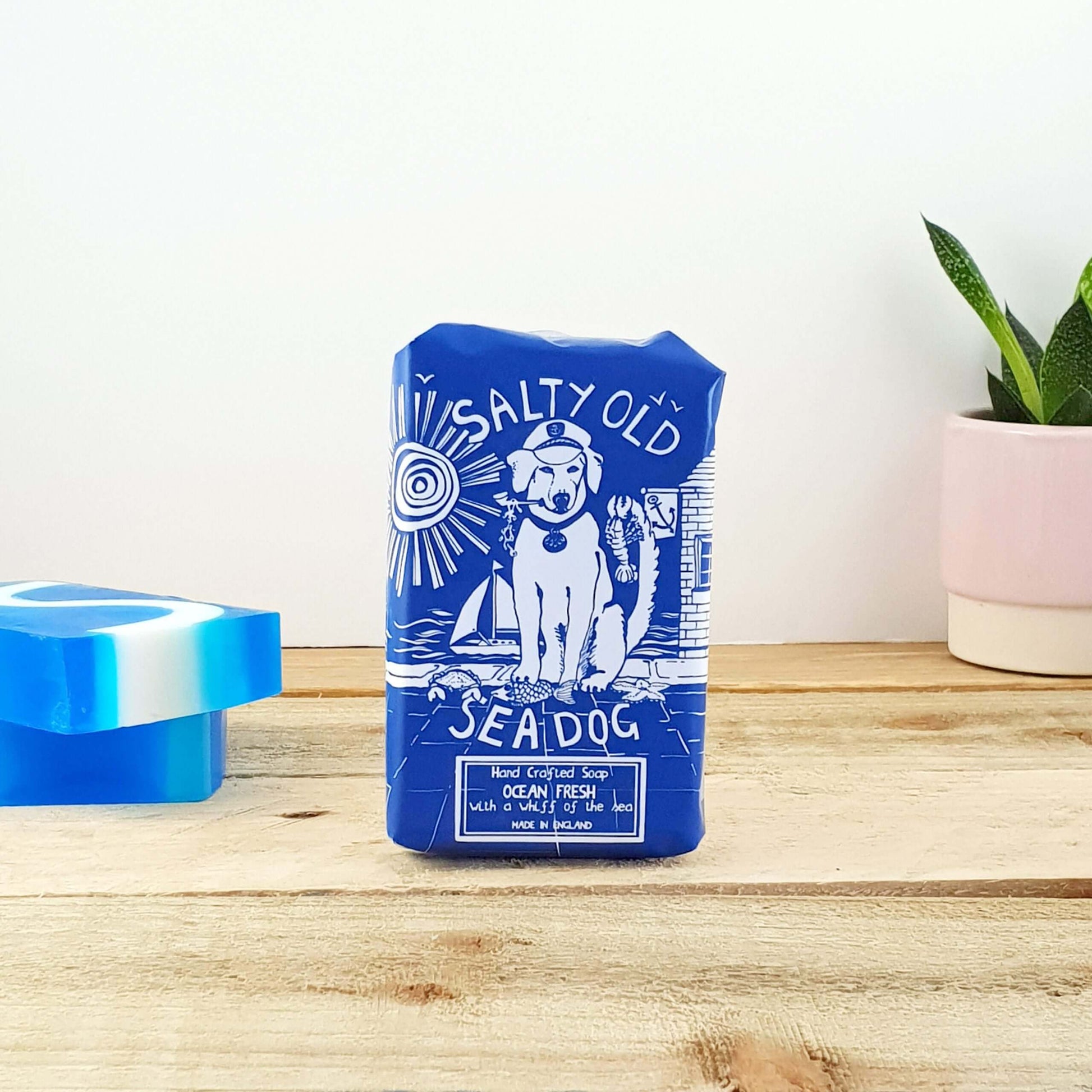 Blue package labeled "Salty Sea Dog Fresh Soap" with a nautical-themed illustration, placed on a wooden surface next to a plant and blue soap