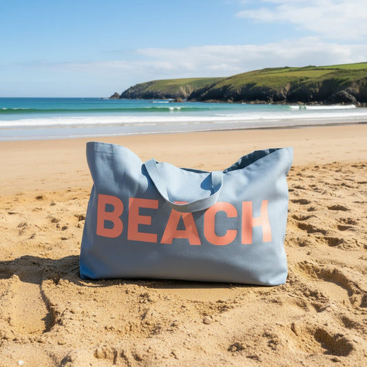 A dusty blue canvas tote bag with the word 'BEACH' printed across the front in a coral pink colour.