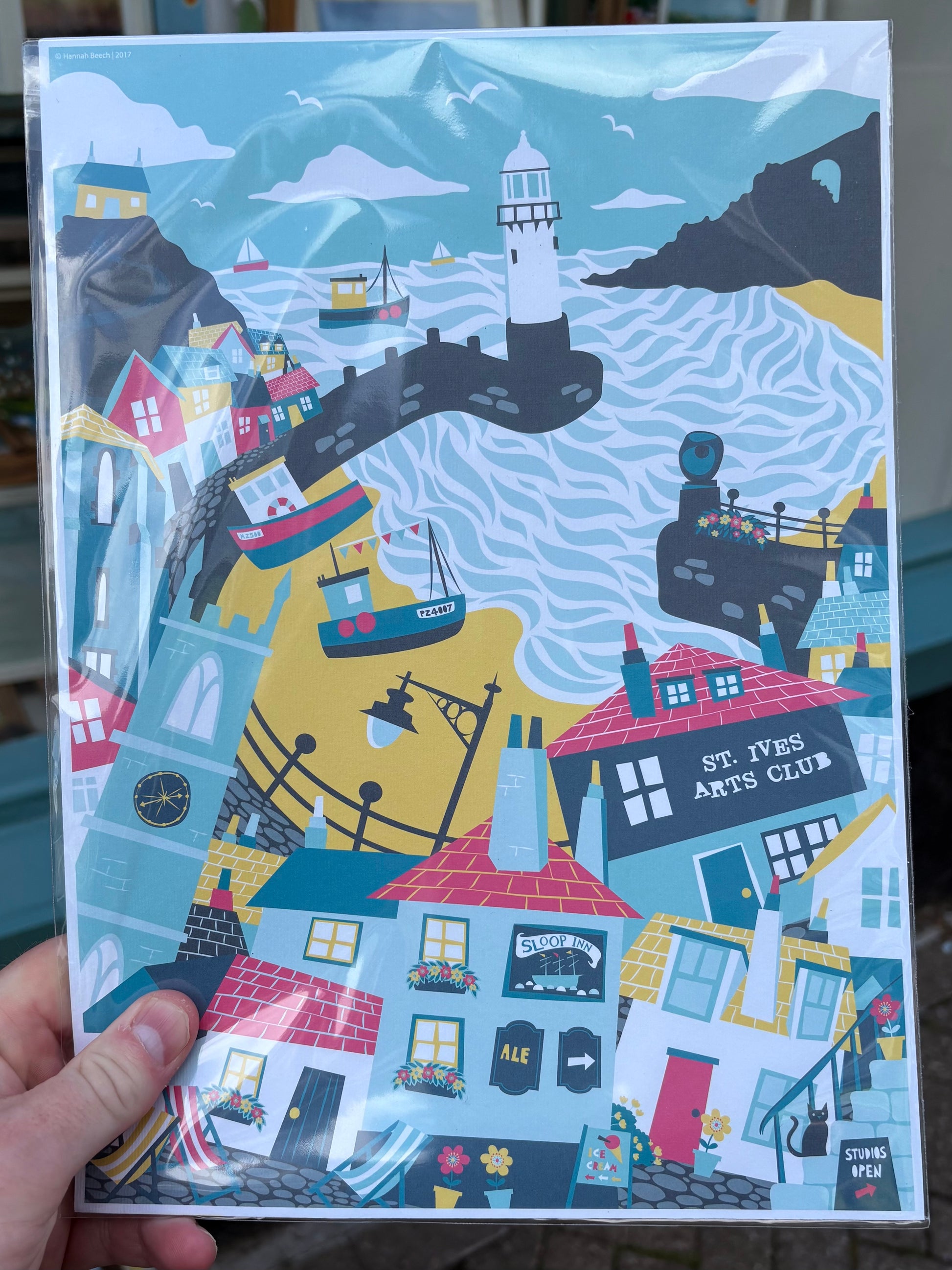 A hand holds a glossy art print featuring a whimsical coastal village scene with colorful houses, boats, a lighthouse, and the text 'ST. IVES ARTS CLUB' on the print.