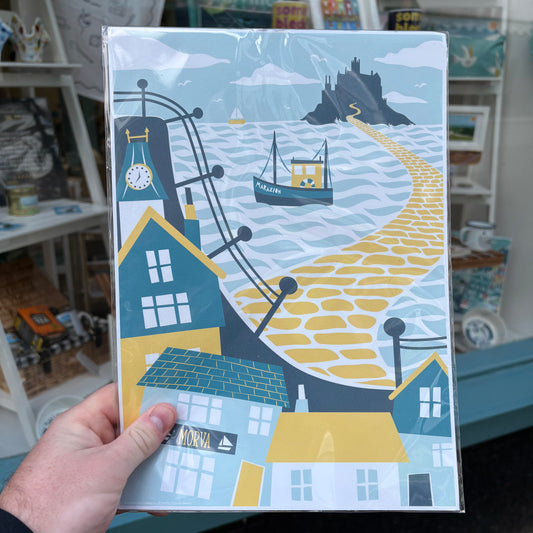 A hand holds a square art print featuring a stylized coastal village scene with a yellow path leading to a rock, blue houses, boats, and ocean waves. 