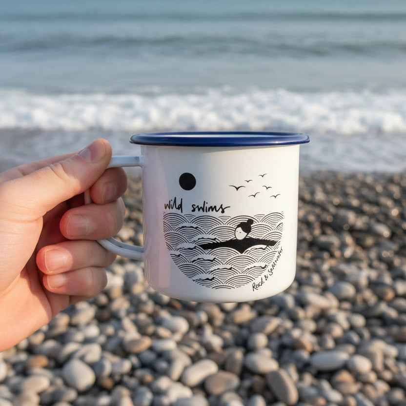 Persons hand holding a mug with a wild swimming design on the side on a beach. The mug has a blue design around the lip.
